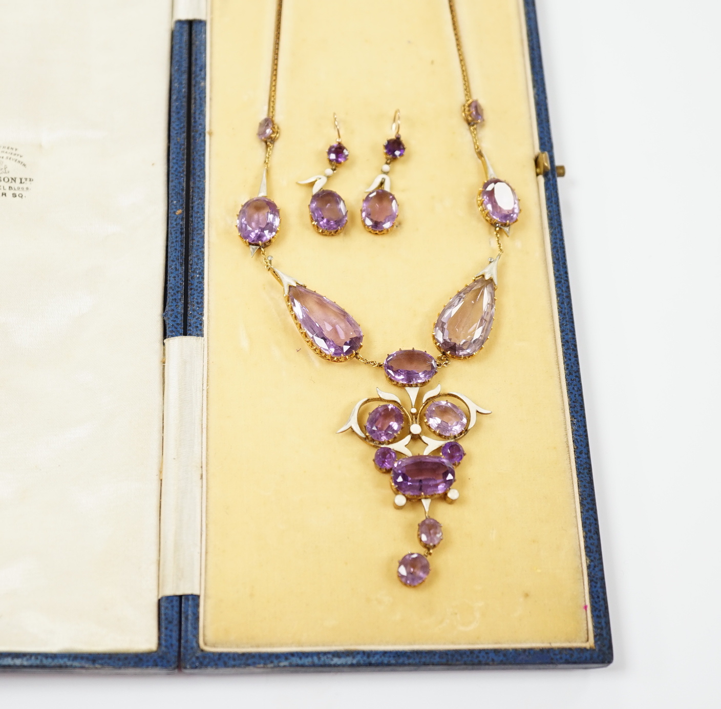 An early 20th century yellow metal, amethyst and enamel set suite of jewellery, comprising a drop necklace, 56cm and a par of matching drop earrings, 33mm, gross weight 38.1 grams.